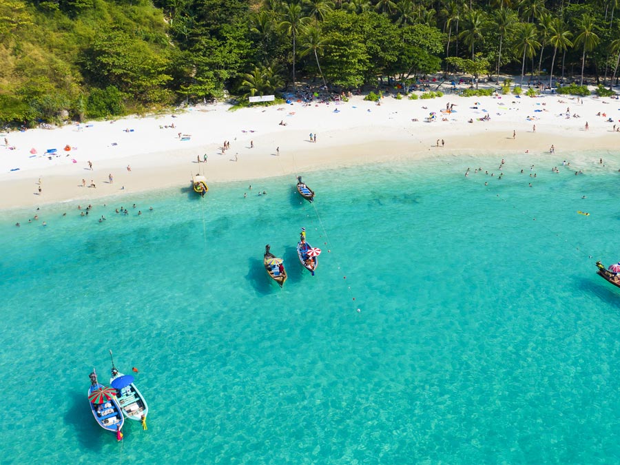 An aerial view of a beautiful tropical beach with white sand and turquoise clear water, longtail boats and people sunbathing, Freedom beach, Phuket, Thailand.